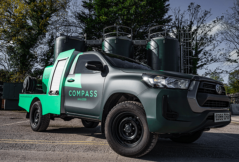 Green and black utility vehicle with the Compass logo parked near fuel storage tanks.