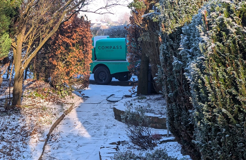 Green fuel delivery vehicle from Compass navigating a snowy garden path surrounded by hedges.
